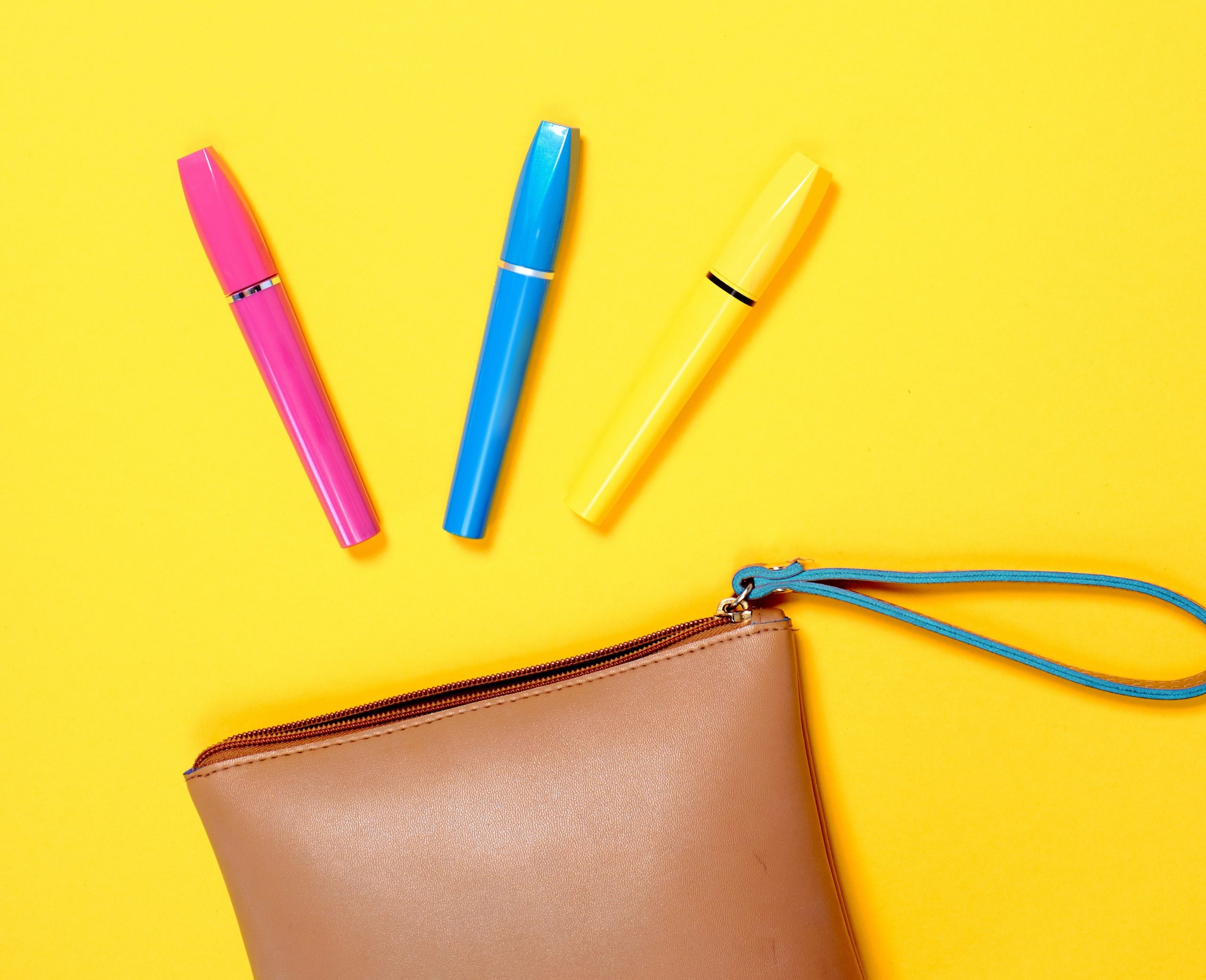 three mascara tubes (pink,blue,yellow) spilling out of small tan purse on yellow plain backdrop