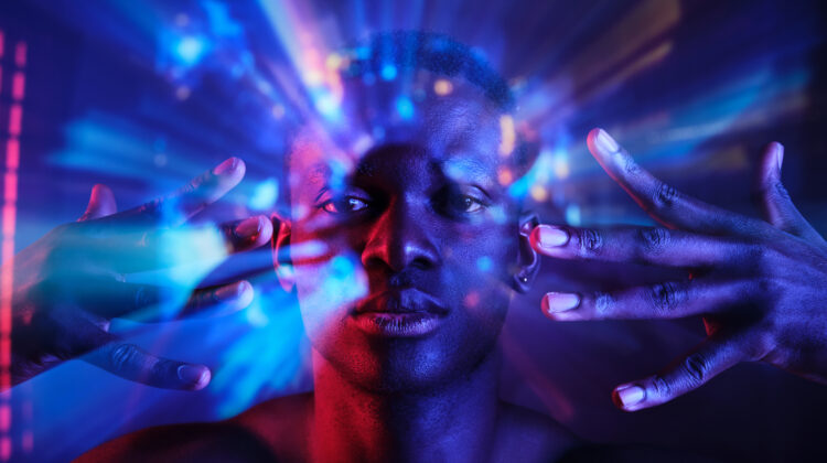 Unemotional young African American male with naked torso holding hands near face and looking at camera while standing in dark neon lights